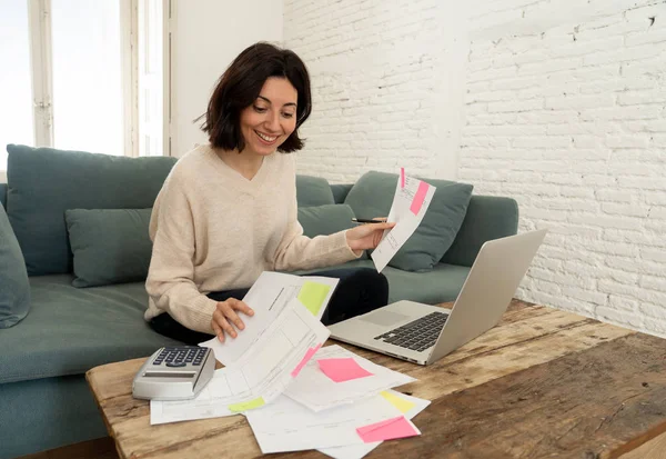 Happy young woman sitting on sofa surrounded by papers calculating expenses and paying bills — Stock Photo, Image