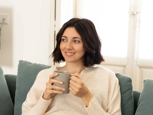 Feliz joven sentada en el sofá en casa con una bebida caliente. En concepto de ocio y tiempo libre —  Fotos de Stock