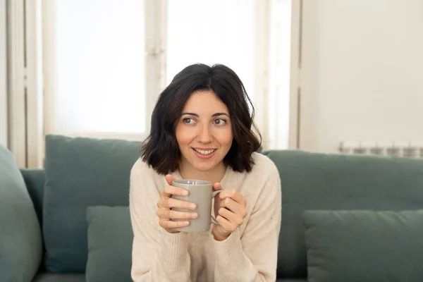 Lifestyle portrait of young pretty relaxed woman drinking hot coffee or chocolate feeling happy and cozy at home smiling happy on the couch. In leisure, peaceful life, happiness lifestyle concept.