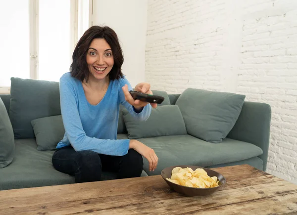 Портрет веселої молодої жінки, що сидить на дивані, дивиться телевізор, що тримає пульт дистанційного керування Ліцензійні Стокові Зображення