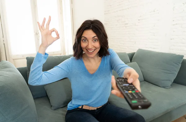 Портрет веселої молодої жінки, що сидить на дивані, дивиться телевізор, що тримає пульт дистанційного керування Стокове Фото