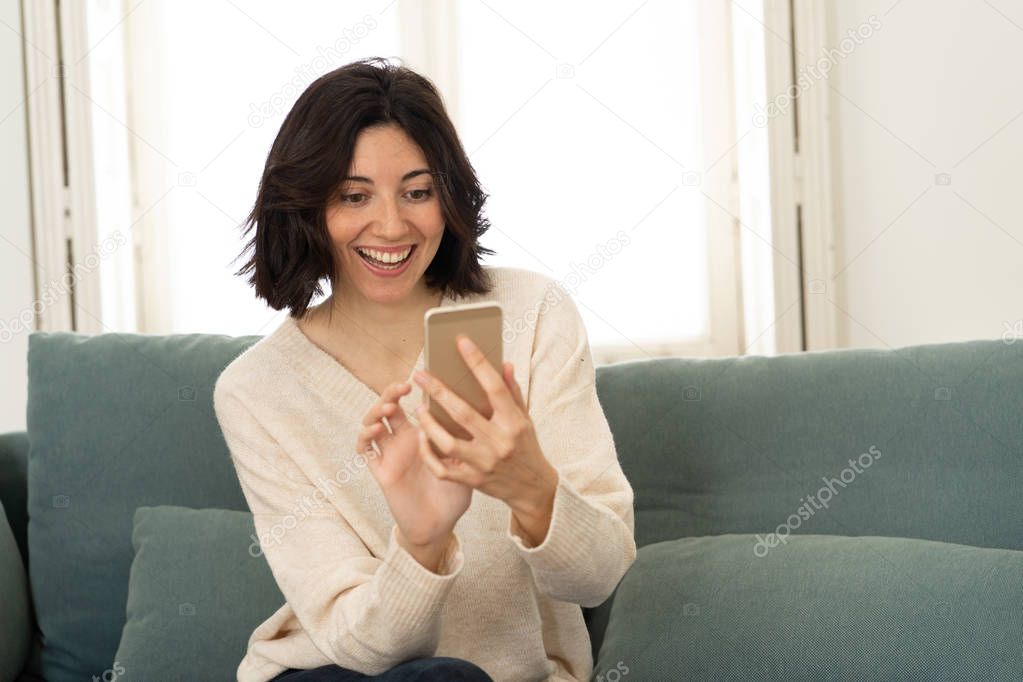 Happy young woman using her smart phone sitting on sofa at home. In leisure and mobile addiction