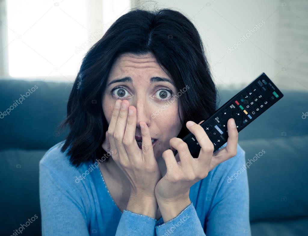 Lifestyle portrait of woman feeling scared and shocked making fear, anxiety gestures while watching horror movie on TV holding remote control. In horror and violence on TV and internet and mass media.