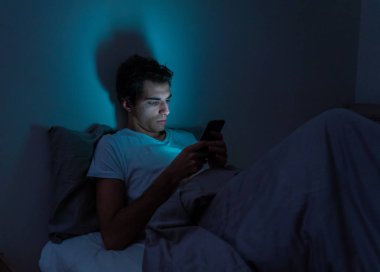 Addicted Man sohbet ve yatakta gece geç saatlere kadar akıllı telefon ile Internette sörf. Sıkılmış, uykusuz ve kasvetli ışık ile karanlık odada yorgun. Uykusuzluk ve mobil bağımlılık kavramı.