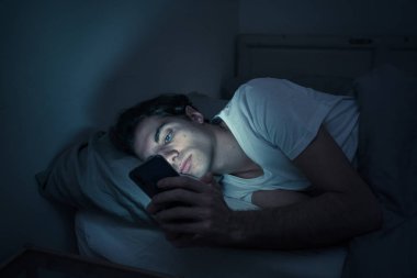 Addicted Man sohbet ve yatakta gece geç saatlere kadar akıllı telefon ile Internette sörf. Sıkılmış, uykusuz ve kasvetli ışık ile karanlık odada yorgun. Uykusuzluk ve mobil bağımlılık kavramı.