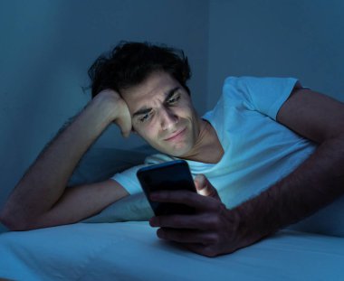 Addicted Man sohbet ve yatakta gece geç saatlere kadar akıllı telefon ile Internette sörf. Sıkılmış, uykusuz ve kasvetli ışık ile karanlık odada yorgun. Uykusuzluk ve mobil bağımlılık kavramı.