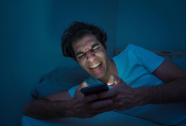 Addicted Man sohbet ve yatakta gece geç saatlere kadar akıllı telefon ile Internette sörf. Sıkılmış, uykusuz ve kasvetli ışık ile karanlık odada yorgun. Uykusuzluk ve mobil bağımlılık kavramı.