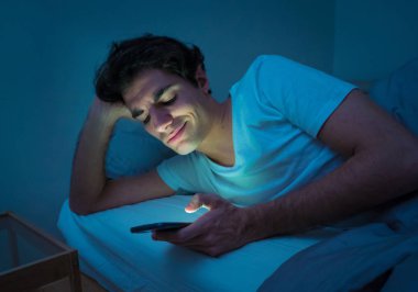 Addicted Man sohbet ve yatakta gece geç saatlere kadar akıllı telefon ile Internette sörf. Sıkılmış, uykusuz ve kasvetli ışık ile karanlık odada yorgun. Uykusuzluk ve mobil bağımlılık kavramı.