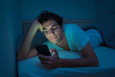 Sosyal medya genç adam sohbet ve yatakta gece akıllı telefon Internette sörf Addicted. Mobil ekran ışığı ile karanlık yatak odasında uykusuz. Uykusuzluk ve çevrimiçi ağ bağımlılığı.