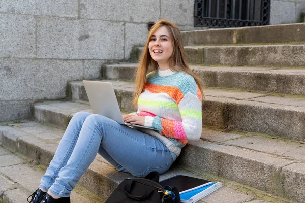 Attractive trendy teen student or young college girl working on laptop on the internet, blogging, chatting and checking email on steps in a european city. In Student learning online City lifestyle.