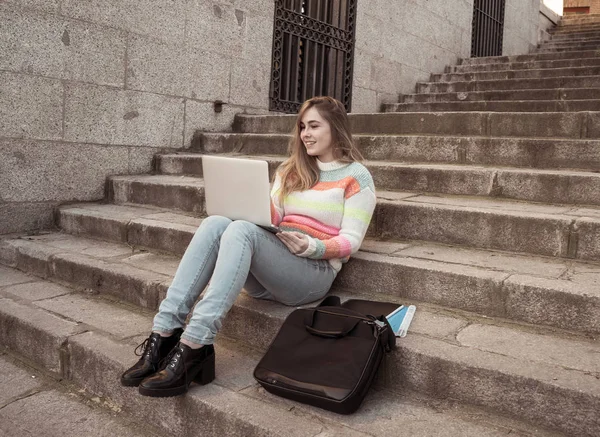 Attractive trendy teen student girl or young college woman working on laptop on the Internet, blogging, chatting or on steps in urban background In Online learning and Social media trends.