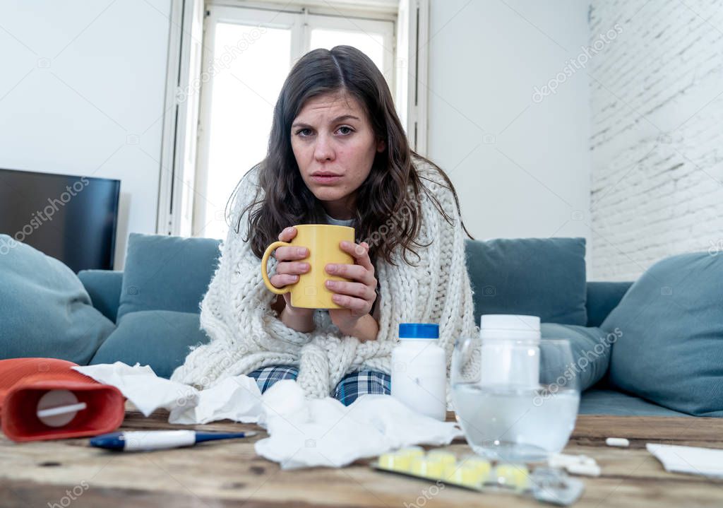 Sick young woman sitting on sofa drinking hot drink cold flu medicine feeling unwell with headache sore nose and high temperature not being able to go to work. Health care and medical concept.