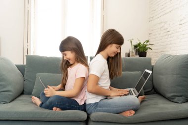Tablet ve dizüstü bilgisayar ile ablası ile güzel sevimli küçük kız birlikte gülümseyerek oynarken ve evde kanepede eğlenmek. Mutlu aile, dijital teknoloji, yaşam tarzı konsepti.