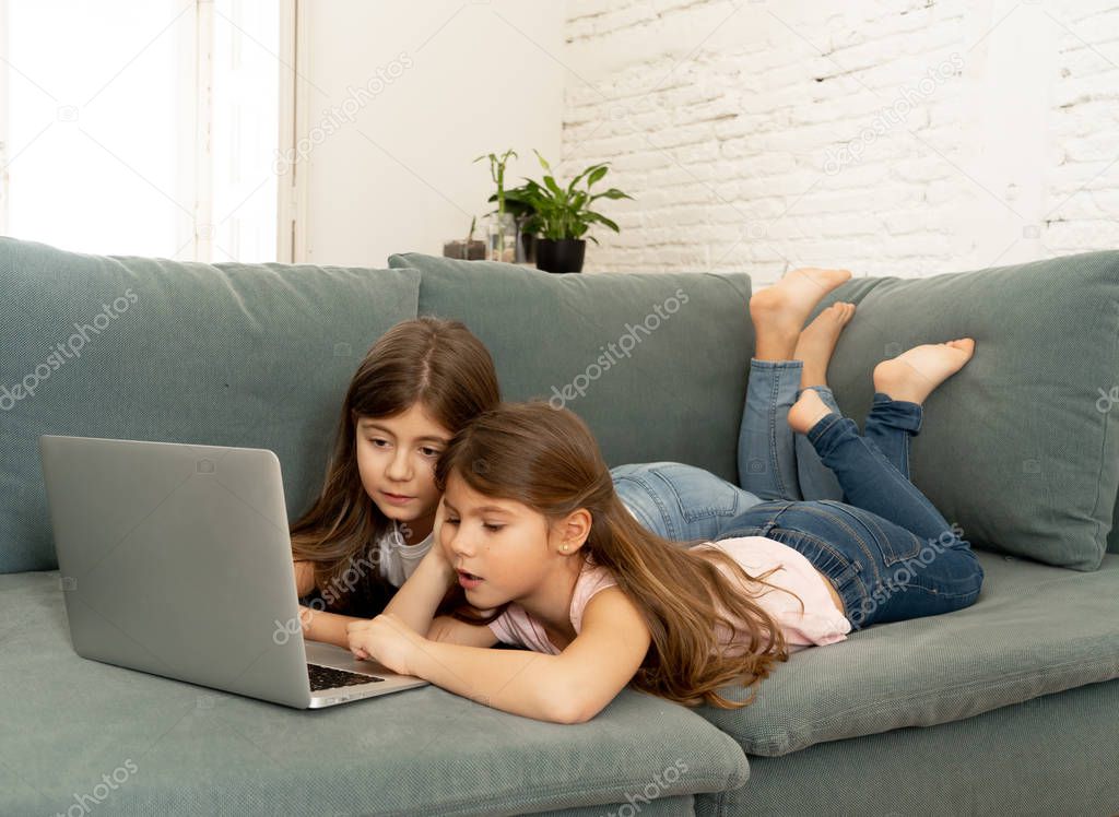 Cute little girl and older sister playing together smiling and having bonding time using a laptop on couch at home. Happy family Siblings relationship and digital technology lifestyle concept.