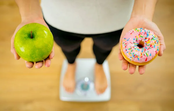 Close up of woman on scale holding on hands apple and doughnut making choice between healthy unhealthy food dessert while measuring body weight in Nutrition Health care Diet and temptation concept.