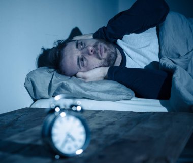 Uykusuz ve çaresiz genç beyaz adam Uykusuz geceleri uyku mümkün değil, hayal kırıklığına ve endişe saat uykusuzluk stres ve uyku bozukluğu kavramı muzdarip bakıyor.