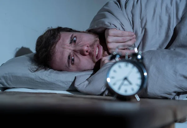 Mental health, Insomnia and sleeping disorders. Frustrated and hopeless sleepless man looking in distress at alarm clock awake at night not able to sleep suffering anxiety caused by stress at work.