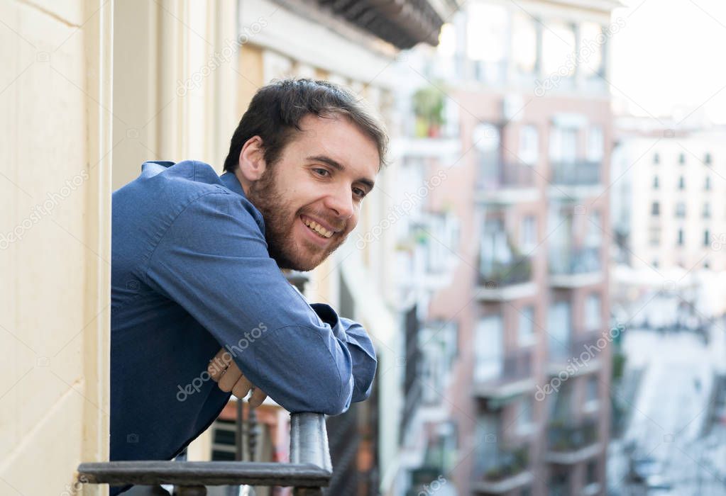 Happy good looking bearded man smiling enjoying urban view on outdoor balcony. Happy young hipster in European city relaxing on room balcony. Home comfort lifestyle and traveling around Europe.