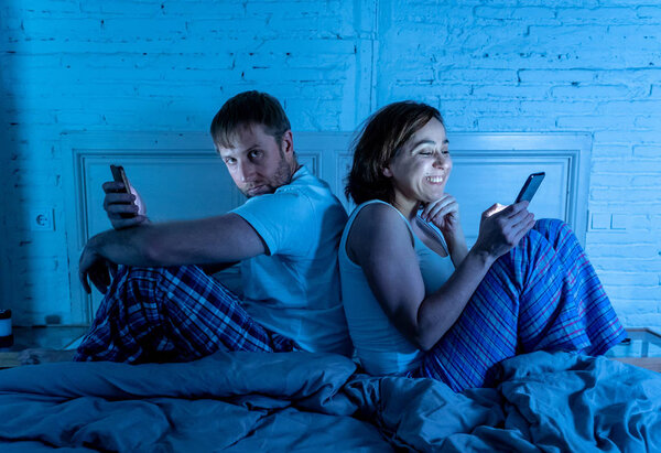 Lifestyle portrait of happy girlfriend wife addicted to mobile phone in bed and angry upset frustrated husband boyfriend bored on phone feeling ignored. Internet mobile addiction and Couple problems.
