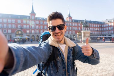 Plaza Mayor, Madrid, İspanya akıllı telefon ile gülümseyen bir selfie çekerek mutlu ve heyecanlı eğlenceli sırt çantası ile Çekici genç turist öğrenci adam. Tatil ve dünya konsepti etrafında seyahat.
