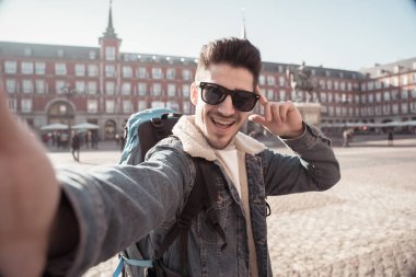 Çekici genç turist öğrenci adam mutlu ve heyecanlı bir komik surat selfie akıllı telefon ile Plaza Mayor, Madrid, İspanya alarak eğlenmeye sırt çantası. Tatil ve seyahat etmek çevrede dünya kavramı.