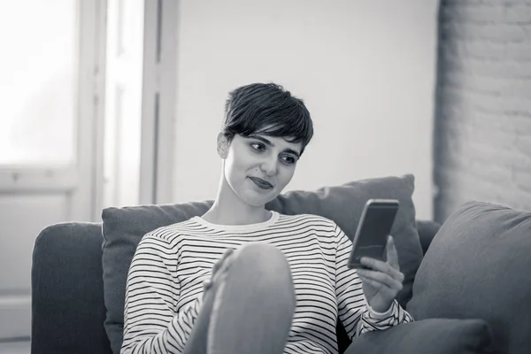 Happy attractive short haired young woman on mobile smart phone chatting and using social media apps sitting on couch at home in dating on line,using new technology and social network concept.