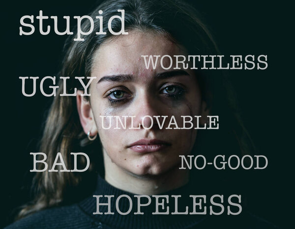 Closeup of sad, depressed schoolgirl victim of bullying on the background and insults written on the foreground isolated on black background and dramatic light. In verbal abuse and harassment.