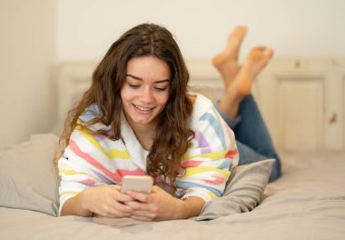 Sosyal medya uygulamaları sohbet veya online kişi, yaşam tarzı, iletişim ve yeni teknoloji kavramı evde yatakta yatan oyun cep telefonuna mesaj gönderme mutlu güzel kız.