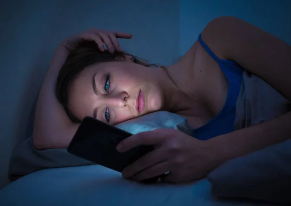 Addicted young beautiful woman in bed, chatting and surfing on the internet using her smart phone sleepy, dull and tired late at night in mobile addiction and technology overuse concept.