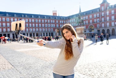 Beautiful young woman happy and excited in Plaza Mayor Madrid Spain taking selfie or video with selfie stick posing for her followers, enjoying historical architecture. In tourism, and travel Europe. clipart