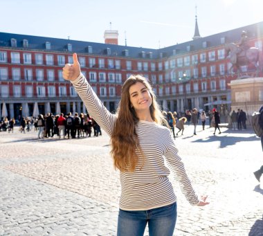 Plaza Mayor Madrid'de mutlu ve heyecanlı genç beyaz bir kadın. Neşeli görünmek, eğlenmek ve güneşli bir günde başparmak yapmak. Turizmde, Avrupa'da seyahat.