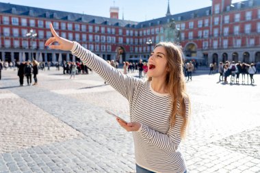 Beautiful young tourist woman happy and excited in Plaza Mayor Madrid Spain. Looking cheerful, checking smart mobile phone pointing at spanish architecture. In tourism travel app and technology. clipart