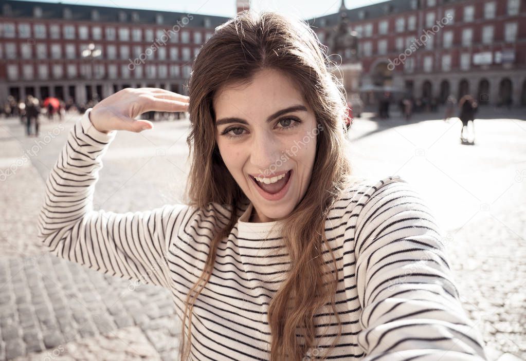 Beautiful young caucasian woman happy and excited in Plaza Mayor Madrid holding the camera and taking a photo of herself. Looking cheerful and joyful. In tourism, European city, travel in Europe.