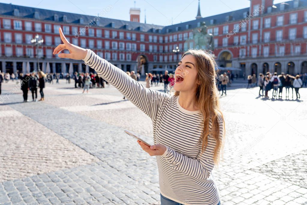 Beautiful young tourist woman happy and excited in Plaza Mayor Madrid Spain. Looking cheerful, checking smart mobile phone pointing at spanish architecture. In tourism travel app and technology.