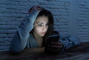 Dramatic portrait of sad scared young woman victim of online harassment and cyberbullying. looking at smart mobile phone stressed and in fear being online abused by stalker. In Dangers of internet. clipart