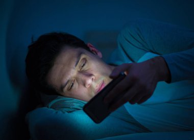 Addicted Man sohbet ve yatakta gece geç saatlere kadar akıllı telefon ile Internette sörf. Sıkılmış, uykusuz ve kasvetli ışık ile karanlık odada yorgun. Uykusuzluk ve mobil bağımlılık kavramı.