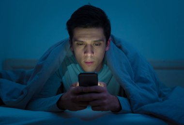 Addicted Man sohbet ve yatakta gece geç saatlere kadar akıllı telefon ile Internette sörf. Sıkılmış, uykusuz ve kasvetli ışık ile karanlık odada yorgun. Uykusuzluk ve mobil bağımlılık kavramı.