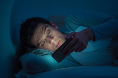 Genç cep telefonu bağımlısı adam uyanık gece yatakta sohbet için akıllı telefon kullanarak, flört ve metin mesajı gönderme. İletişim, Internet bağımlılığı ve sosyal medya ağ istismarı kavramı