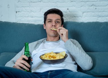 Lifestyle portrait of young bored man on couch with remote control zapping for movie or live sport. Looking disinterested drinking beer. Sedentary and mass social media or Television addiction. clipart