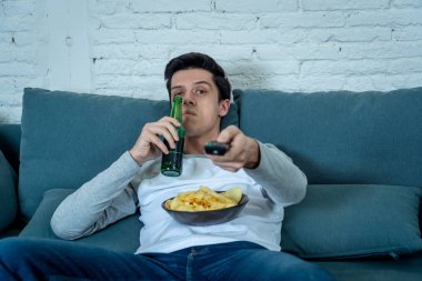 Film veya canlı spor için uzaktan kumanda zap ile kanepede genç sıkılmış adamın Lifestyle portre. İlgilendiğim bira içmeye bakıyor. Sedanter ve kitle sosyal medya veya televizyon bağımlılığı.