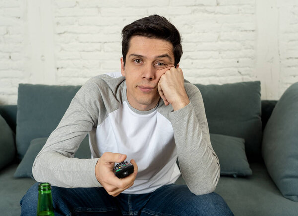 Lifestyle portrait of young bored man on couch with remote control zapping for movie or live sport. Looking disinterested drinking beer. Sedentary and mass social media or Television addiction.
