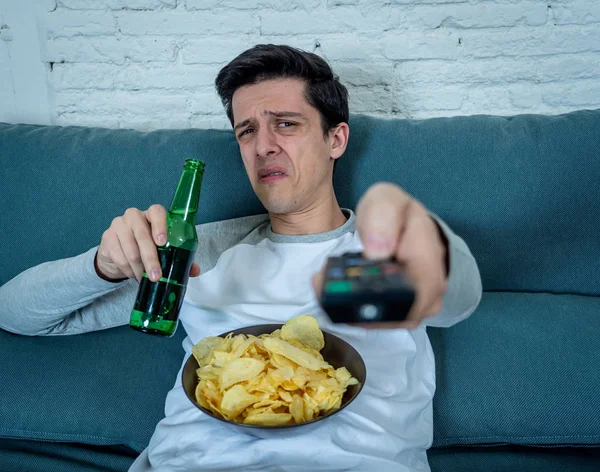 Young man switching channels in dislike feeling disgust with TV shows or movies. With a look of disapproval while drinking beer. Rubbish in and bad television shows and unhealthy lifestyle.