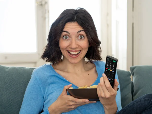 Happy woman on sofa with TV remote control ready to watch favorite movie of TV Show. Looking enthusiastic, making gestures of approval and eating chips. In people, technology and leisure concept.