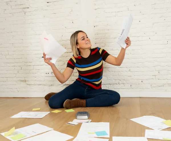 Happy young woman sitting on the floor surrounded by papers calculating costs, charges, taxes or paying bills at new home. In home finances, mortgage, loan banking and house investment concept.