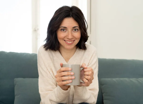 Lifestyle portrait of young pretty relaxed woman drinking hot coffee or chocolate feeling happy and cozy at home smiling happy on the couch. In leisure, peaceful life, happiness lifestyle concept.