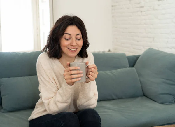 Lifestyle portrait of young pretty relaxed woman drinking hot coffee or chocolate feeling happy and cozy at home smiling happy on the couch. In leisure, peaceful life, happiness lifestyle concept.