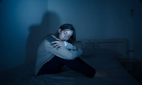 Young teenager girl suffering mobile cell phone addiction feeling lonely and depressed having insomnia needing to be connected sitting on bed late at night. In