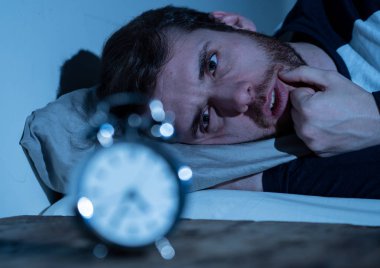 Uykusuz ve çaresiz genç beyaz adam Uykusuz geceleri uyku mümkün değil, hayal kırıklığına ve endişe saat uykusuzluk stres ve uyku bozukluğu kavramı muzdarip bakıyor.
