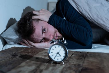 Ruh sağlığı, uykusuzluk ve uyku bozuklukları. Sinirli ve umutsuz uykusuz adam alarm saat uyanık gece uyanma anksiyete İşteki stres neden acı uyku mümkün değil bakıyor.