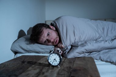 Ruh sağlığı, uykusuzluk ve uyku bozuklukları. Sinirli ve umutsuz uykusuz adam alarm saat uyanık gece uyanma anksiyete İşteki stres neden acı uyku mümkün değil bakıyor.